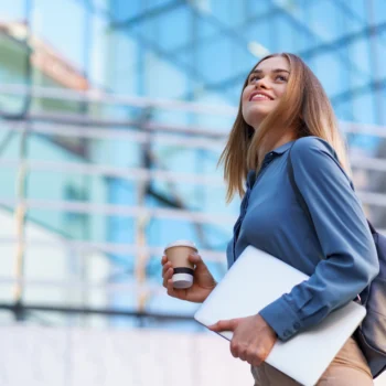Mulher sorrindo ao ar livre, segurando um notebook e um copo de café, em frente a um edifício corporativo moderno.